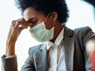 Black businesswoman with protective face mask holding her head in pain.
