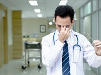 Asian senior doctor man wearing medical uniform standing while take off glasses with tired rubbing nose and eyes feeling fatigue and headache in hospital corridor. Stress and frustration concept.