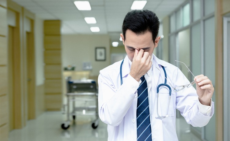 Asian senior doctor man wearing medical uniform standing while take off glasses with tired rubbing nose and eyes feeling fatigue and headache in hospital corridor. Stress and frustration concept.