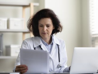 Focused serious middle aged GP doctor reading medical history, patient records, Chief physician reviewing insurance agreement, contract, paper documents at workplace with laptop. Medical job concept