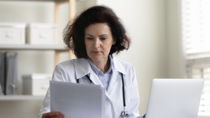 Focused serious middle aged GP doctor reading medical history, patient records, Chief physician reviewing insurance agreement, contract, paper documents at workplace with laptop. Medical job concept