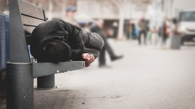 Poor homeless man or refugee sleeping on the wooden bench on the urban street in the city, social documentary concept, selective focus