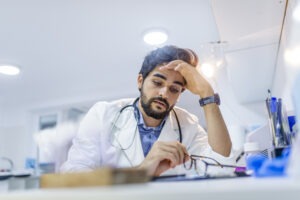 Overworked doctor in his office. Tired male scientist trying to focus, rubbing his forehead with fingers. Mid adult male doctor working long hours. Stressed male doctor sat at his desk
