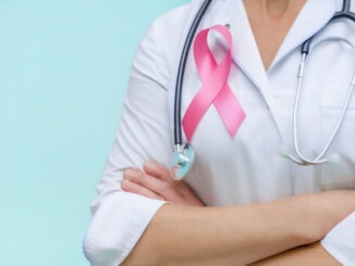 NEWS: New Targeted Breast Cancer Drug Approved in UK Doctor with folded arms and a stethoscope on his neck, shows a pink ribbon close-up on uniform on a blue background.