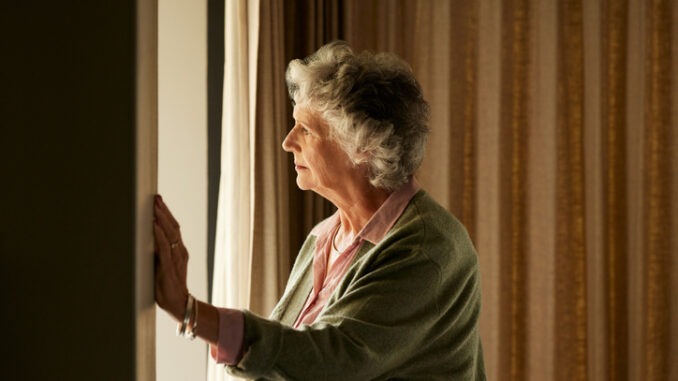 Thoughtful senior woman standing near a window at home feeling lonely. Elder woman looking outside window with uncertainty while standing indoors with copy space. Doubtful old lady gazing throught a window in a dimly lit room.