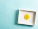 Concept of well-being, well done, feedback, employee recognition award. Happy yellow smiling emoticon face frame hanging on blue background with empty space for text.