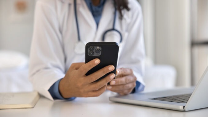 Close up female doctor using smartphone at workplace, access records, review medical histories, conduct virtual consultations with patients, provide advice, prescribe medication through telehealth app